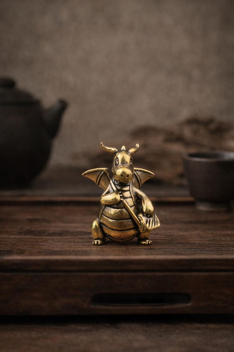 May include: A small, metallic brass-colored figurine of a dragon with wings, horns, and a rounded belly. The dragon is depicted in a standing position, holding a long, thin object in its hands, featuring a textured surface and a warm, golden finish.