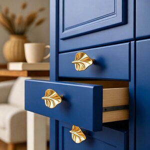 May include: A deep blue cabinet featuring three gold-colored metal drawer pulls shaped like leaves. The middle drawer is partially open, revealing a light-colored wooden interior. The cabinet has a smooth, matte finish and clean, straight lines.