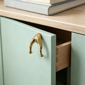 Peut inclure: Une poign&eacute;e de placard en m&eacute;tal dor&eacute; poli en forme de fourchette, mont&eacute;e sur une porte de placard vert menthe clair. La poign&eacute;e pr&eacute;sente une surface lisse et r&eacute;fl&eacute;chissante ainsi qu'un design incurv&eacute; et bifurqu&eacute; qui s'&eacute;tend vers l'ext&eacute;rieur.