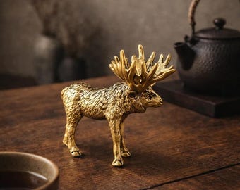 May include: A gold-colored metal figurine of a standing moose with detailed fur texture and large, branching antlers. The moose is depicted in a side profile view, showcasing its muscular build and distinct facial features in a metallic finish.
