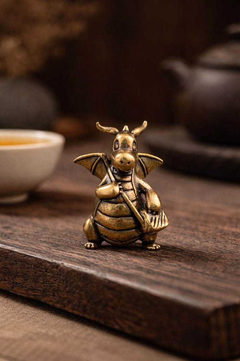 May include: A small, detailed brass dragon figurine with wings, horns, and a satchel. The dragon is posed on a wooden surface, with a cup and teapot in the background. The figurine is approximately 2 inches tall.