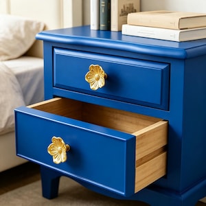 May include: A vibrant blue nightstand featuring two drawers with gold-toned, flower-shaped metal knobs. The top drawer is closed, while the bottom drawer is partially open, revealing a light-colored wooden interior structure and smooth finish.