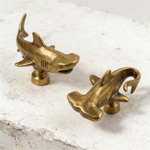 May include: Two brass-colored metal cabinet knobs shaped like hammerhead sharks. Each knob features a detailed shark figure with a wide, flat head, dorsal fin, and side fins, mounted on a small cylindrical base for attachment to furniture.
