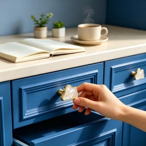 May include: A cloud-shaped cabinet knob with a white, pearlescent inlay and a gold-toned metal base. The knob is attached to a deep blue drawer front with a recessed panel design. A second identical knob is visible on the adjacent drawer.