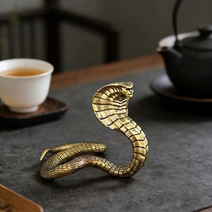 Könnte beinhalten: Eine detaillierte Cobra-Skulptur aus Messing mit erhobenem Kopf und strukturierter Schuppung. Die Schlange befindet sich auf einer dunklen Oberfläche, im Hintergrund eine Teetasse und eine Teekanne. Die Skulptur ist etwa 15 cm lang.