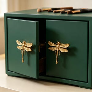 May include: A dark green storage cabinet with two doors, each featuring a gold-colored dragonfly-shaped handle. The top of the cabinet holds several makeup brushes with brown bristles and black handles, arranged in a horizontal row.