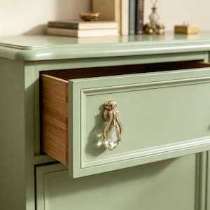 May include: A sage green wooden chest of drawers featuring a single partially open drawer. The drawer front is adorned with a decorative gold-toned metal pull handle that incorporates a clear, faceted, teardrop-shaped crystal accent in the centre.