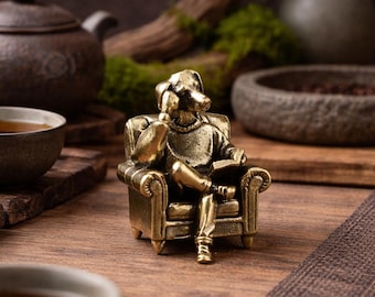May include: A small, detailed brass figurine of a dog seated in a miniature armchair, reading a book. The dog is wearing a sweater and appears to be deep in thought. The figurine is a unique decorative item, perfect for a desk or shelf.