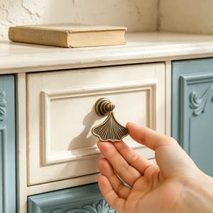 May include: A brass-toned metal drawer pull shaped like a ginkgo leaf with a fan-like design and vertical ridges. The hardware is attached to a cream-colored wooden drawer front with a rectangular frame detail on a piece of furniture.