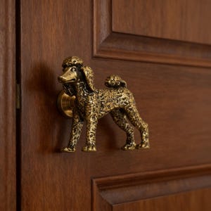 Puede incluir: Un pomo de puerta de metal en tono latón con forma de perro caniche de pie. La figura presenta un acabado texturizado que imita un pelaje rizado y está montada horizontalmente en un panel de puerta de madera oscura como manija funcional.