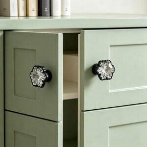 May include: Two cabinet knobs featuring a clear, faceted, flower-shaped crystal center set within a black, scalloped-edge frame. These decorative hardware pieces are mounted on the light sage green doors of a cabinet with a recessed panel design.