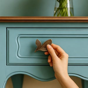 Peut inclure: Une poignée de tiroir en métal couleur bronze en forme de feuille stylisée avec des textures de nervures détaillées. La poignée est fixée sur une façade de tiroir en bois bleu clair dotée d'un panneau rectangulaire encastré.