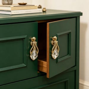 May include: A dark green cabinet featuring two doors with recessed panels and gold-toned metal drop handles. Each handle is accented with a clear, faceted crystal-like teardrop pendant. One door is slightly ajar, revealing a natural wood interior.