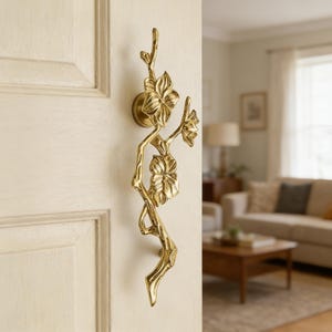 May include: A vertical door handle made of polished gold-toned metal, shaped like a slender tree branch with three detailed flower blossoms attached. The handle is mounted onto a light-colored wooden door, featuring a smooth, reflective surface.