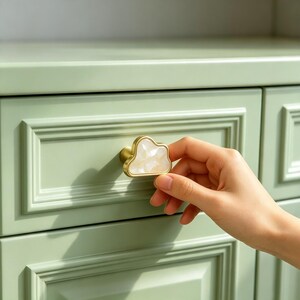 May include: A cloud-shaped cabinet knob featuring a gold-toned metal frame and a white, marbled-effect resin center. The knob is mounted on a light sage green drawer front with a recessed panel design, showcasing a smooth, glossy finish.