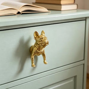 Puede incluir: Un tirador de cajón de metal dorado con forma de bulldog francés. El tirador presenta una escultura tridimensional detallada de la cabeza, el pecho y las patas delanteras del perro, montada en un frente de cajón de madera pintado de verde claro.