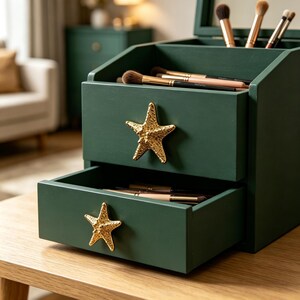May include: A dark green wooden makeup organiser featuring two drawers and an open top compartment. Each drawer is equipped with a textured, gold-coloured starfish-shaped handle. Several makeup brushes are stored inside the drawers and top.