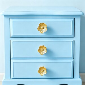 May include: A light blue bedside table featuring three horizontal drawers, each equipped with a single gold-colored metal knob shaped like a five-petaled flower with a textured center. The furniture has a smooth finish and a rectangular frame.