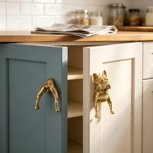 Peut inclure: Deux boutons de placard en métal doré en forme de bouledogue français. Un bouton est fixé sur une porte de placard bleue, montrant l'arrière du chien, et l'autre est fixé sur une porte de placard blanche, montrant l'avant du chien avec des oreilles détaillées.