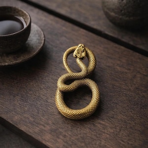Op de afbeelding: Een gouden, slangvormig decoratief object, mogelijk een ring of een presse-papier, ligt op een donker houten oppervlak. De slang is opgerold, met zijn kop omhoog. Een kopje thee en een kleine keramische pot staan op de achtergrond.