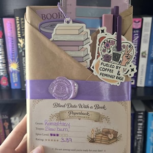 Può includere: Un libro avvolto in carta marrone intitolato Blind Date With a Book, fissato con un nastro viola e un sigillo di ceralacca. Include un adesivo con libri impilati, un adesivo di caffè con il testo Fueled by Coffee & Feminist Rage e dei foglietti adesivi.