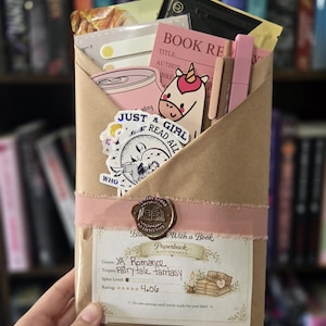 May include: A brown paper package tied with a pink ribbon, containing pink and white items, such as stickers, a pink can, and a book review card. The package is sealed with a wax stamp and labelled "With a Book".