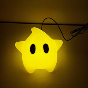 Pode incluir: Uma lâmpada amarela brilhante em forma de estrela com uma superfície lisa e luminosa. A lâmpada apresenta dois grandes olhos ovais pretos sólidos posicionados centralmente e um pequeno detalhe circular no topo. Um fino cabo de alimentação preto.