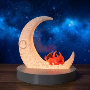 Può includere: Una lampada a forma di mezzaluna con superficie testurizzata poggiata su una base circolare grigia. Una piccola figura di drago arancione alato è raggomitolata e dorme sulla curva inferiore della luna, che emette una luce calda e soffusa.