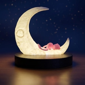 Peut inclure: Une lampe en forme de croissant de lune à la surface texturée blanc cassé, reposant sur une base circulaire noire. Une petite créature rose, semblable à un félin, est recroquevillée sur la courbe intérieure de la lune illuminée et douce.