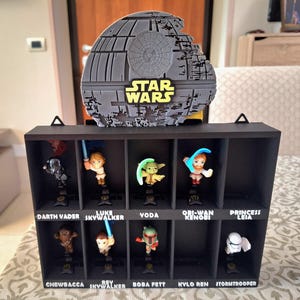 Könnte beinhalten: Ein schwarzes Display-Regal mit Star Wars-Charakterfiguren. Das Regal hat ein graues Todesstern-Design und die Worte "STAR WARS". Die Figuren umfassen Darth Vader, Luke Skywalker, Yoda und andere. Jede Figur ist mit ihrem Charakternamen beschriftet.