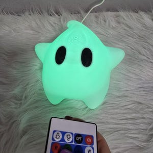 Puede incluir: Una luz de noche brillante en forma de estrella, con ojos negros y una base redondeada. La luz emite un suave brillo verde. Se sostiene un mando a distancia cerca, con botones para ajustar el color y el brillo. La luz de noche está conectada a un cable blanco.
