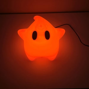 Può includere: Una luce notturna a forma di stella arancione con due occhi ovali neri e una bocca piccola. La luce è collegata con un cavo nero. La luce è luminosa.