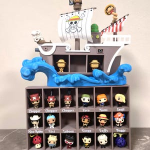 Könnte beinhalten: Ein brauner Kunststoff-Displayständer in Schiffsform mit einem weißen Segel, das ein Totenkopf-Logo zeigt. Die Basis ist ein Gitter mit achtzehn Fächern, die jeweils mit Namen wie Chopper, Zoro, Nami, Sanji, Nico Robin, Vivi, Laboon, Usopp, Luffy, Buggy, Wapol, Mr. 5, Crocodile, Smoker, Hito Hito No Mi und Gomu Gomu No Mi beschriftet sind und kleine Figuren enthalten.