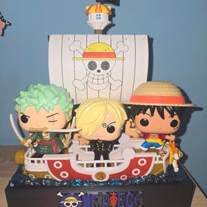 Può includere: Un colorato oggetto da collezione di One Piece con tre personaggi su una base a forma di nave. La nave ha una vela bianca con un emblema a cappello di paglia, una bandiera nera e il testo "ONE PIECE". Le figure includono Zoro, Sanji e Luffy.