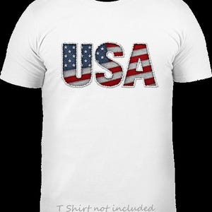 May include: A white short-sleeved t-shirt featuring the text USA in a bold, block font across the chest. The letters are filled with a graphic pattern of the American flag, displaying red and white stripes and a blue field with white stars.