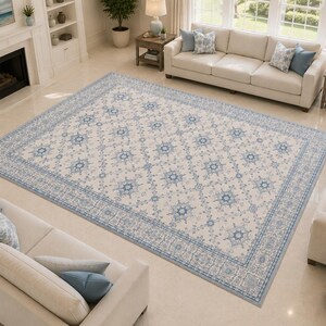 K&ouml;nnte beinhalten: Ein gro&szlig;er rechteckiger Teppich mit cremefarbener Basis, verziert mit einem sich wiederholenden Muster aus blauen Blumenmedaillons und komplexen geometrischen Bord&uuml;ren. Der Teppich liegt mittig in einem Wohnraum mit hellem Boden.