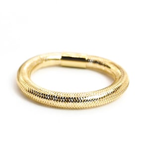 Könnte beinhalten: Ein goldfarbenes Armband mit einem strukturierten, gewebten Design. Das Armband ist kreisförmig und scheint aus Metall zu bestehen. Der Verschluss ist ein einfacher, rechteckiger Goldbarren. Der Schmuck ist vor einem weißen Hintergrund platziert.