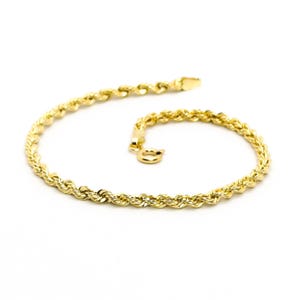 Può includere: Un braccialetto a catena a corda dorata, esposto su sfondo bianco. Il braccialetto è composto da maglie d'oro intrecciate, con chiusura a fermaglio. Il gioiello è un design classico, adatto all'uso quotidiano o alle occasioni speciali.