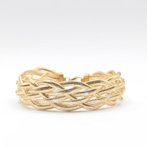 Könnte beinhalten: Ein goldfarbenes Armband aus mehreren Strängen feinen, flexiblen Metalldrahts, die zu einem breiten, geflochtenen Muster verwoben sind. Das Armband hat eine polierte, reflektierende Oberfläche und einen sichtbaren Metallverschluss.