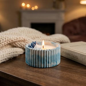 May include: A lit candle in a blue and white striped ceramic container with a white candle and a small succulent arrangement. A thick, cream-colored knitted blanket and a book titled "Evening Tales" are also on the wooden table.