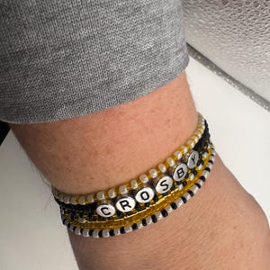 Könnte beinhalten: Ein mehrreihiges Perlenarmband mit einer zentralen Reihe aus silberfarbenen Buchstabenperlen, die das Wort CROSBY bilden. Das Design umfasst abwechselnde Reihen aus kleinen, durchscheinenden weißen, blickdichten schwarzen und gelben Glasperlen.