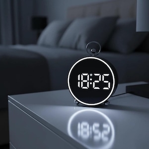 May include: A black digital alarm clock with a white illuminated display showing the time 18:25. The clock is round with a silver handle on top and sits on a white surface. The time is reflected on the surface below.