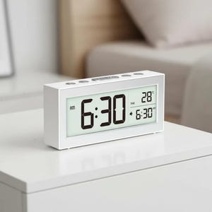 May include: A white digital alarm clock displaying the time as 6:30 AM. The clock also shows the date and temperature. The clock is rectangular and sits on a white surface.