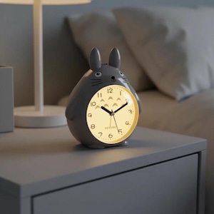 May include: A grey Totoro-shaped alarm clock with a glowing yellow clock face. The clock has black hands, a simple dial, and the word "Totoro" at the bottom. The clock is on a grey bedside table.