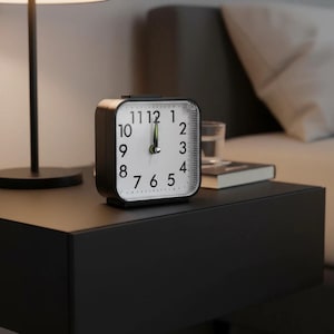 Puede incluir: Un reloj despertador analógico cuadrado en blanco y negro con esfera blanca y números negros. El reloj está sobre una mesita de noche negra. La hora es aproximadamente la 1:00. Una lámpara y una cama están en el fondo.