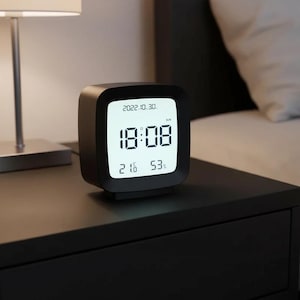 Puede incluir: Un reloj despertador digital negro que muestra la hora, la fecha, la temperatura y la humedad. El reloj muestra las 18:08, la fecha 2022.10.30, la temperatura 21,6 grados Celsius y la humedad 53%. El reloj está sobre una mesita de noche negra.