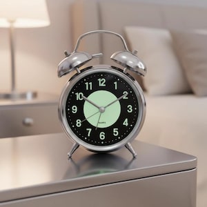 May include: A silver-colored, retro-style alarm clock with a black face and white numerals. The clock has a glowing green center and hands, and is set on a silver nightstand. The clock is a quartz movement.