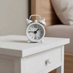 May include: A small, white alarm clock with a classic design sits on a white nightstand. The clock features a round face with black numerals and hands, and two bells on top. The time is approximately 1:10. The nightstand has a single drawer with a round knob.