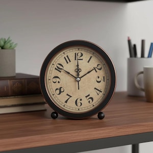 May include: A round, black and bronze-colored analog clock with a cream-colored face and black numerals. The clock has a classic design and sits on a wooden surface. The clock is a decorative item.