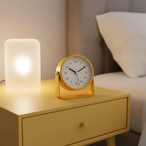 May include: A yellow analog alarm clock with a white face and black hands sits on a yellow nightstand. A white cylindrical lamp is to the left of the clock. The clock is approximately 4 inches wide.
